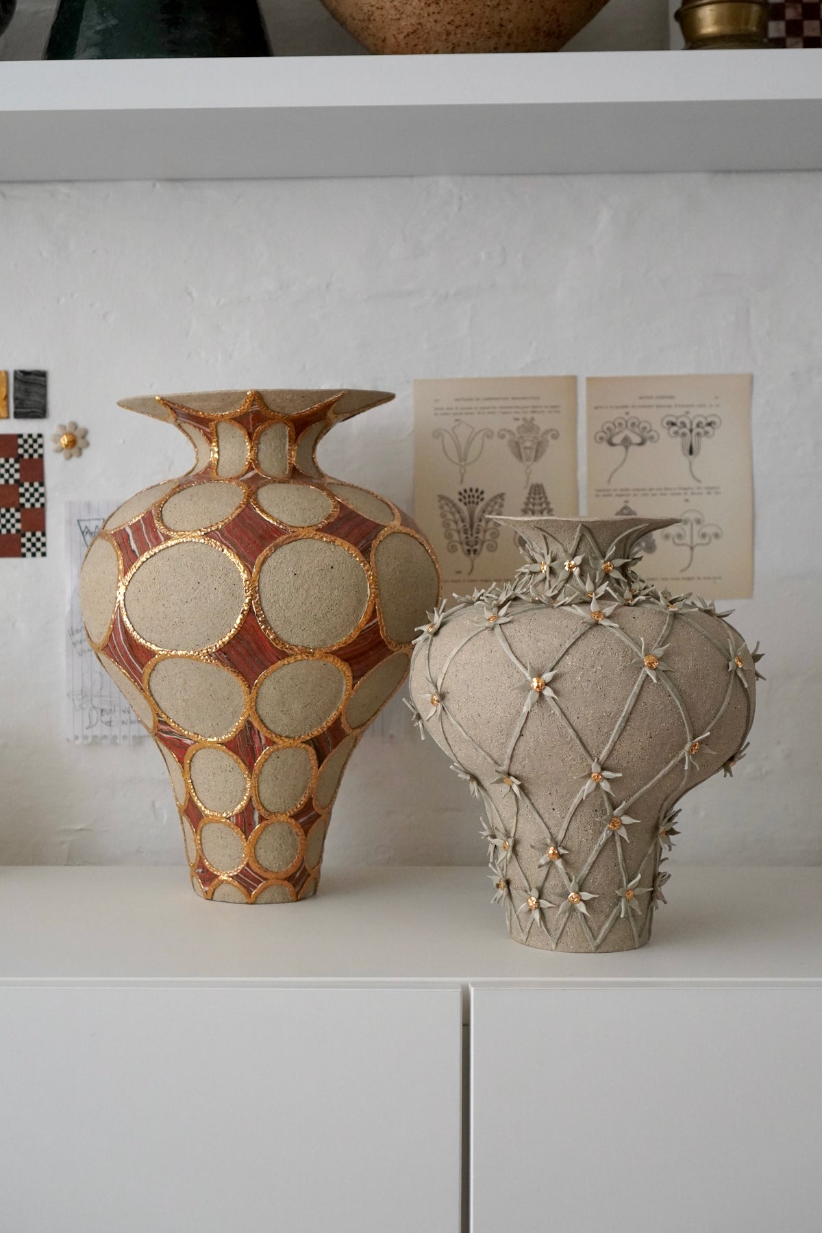 Two decorative ceramic vases displayed on a white shelf against a white wall. The left vase has a warm terracotta orange and beige pattern with gold accents, while the right vase is a textured light beige with a diamond lattice design and small star details.
