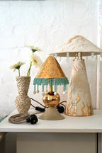 Marley Table Lamp by Tantri Mustika, a pierced gold shade fringed with aqua glass beads, beside a carved white lamp and a vase of daisies