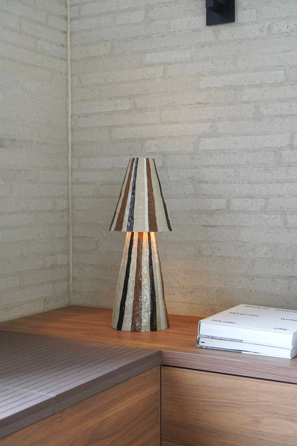 Stellar Table Lamp in Chocolate by Tantri Mustika, lit, on a timber bench against a pale brick wall