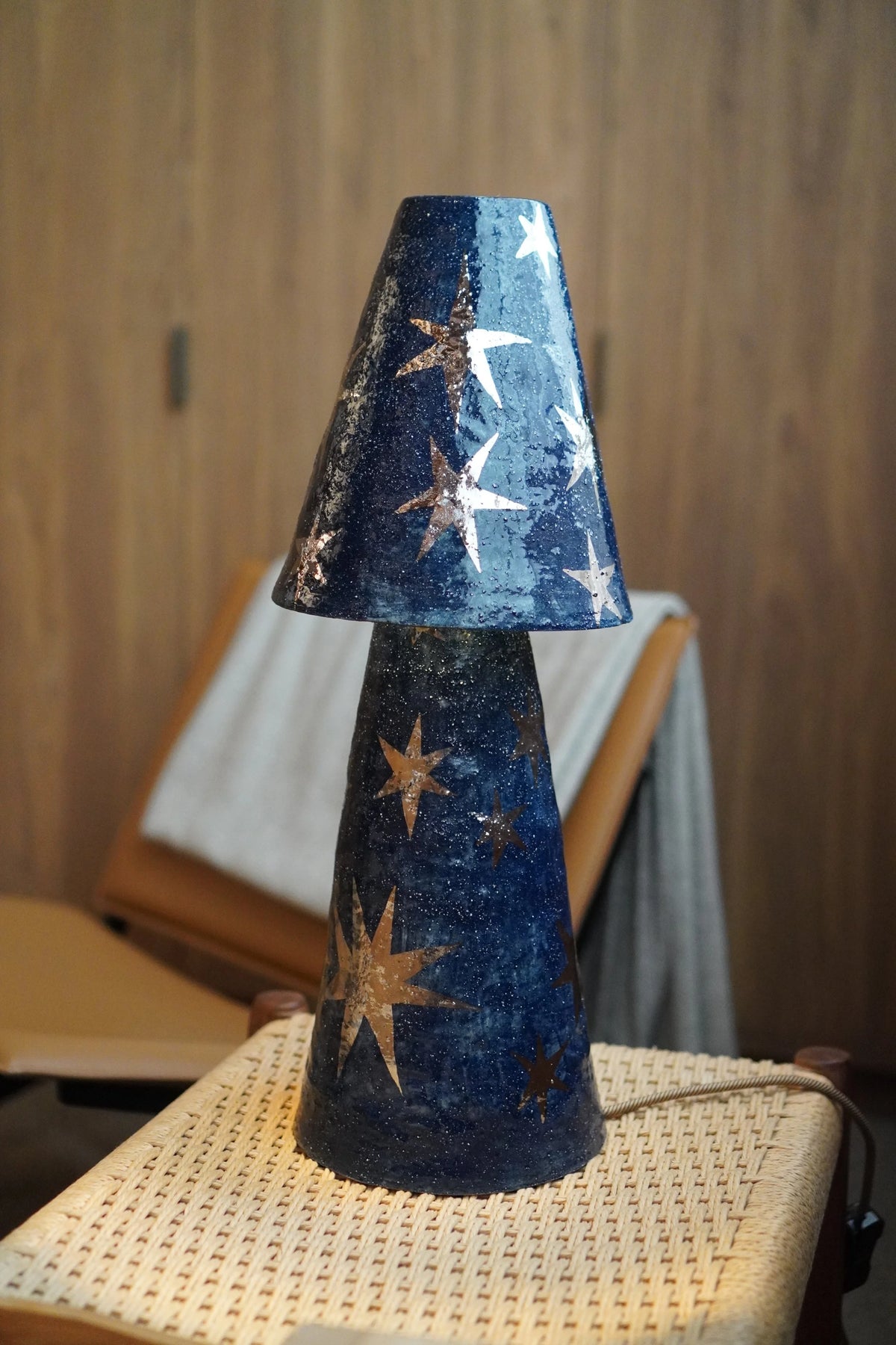 Merlin Table Lamp by Tantri Mustika, deep navy stoneware with platinum lustre stars, on a woven stool