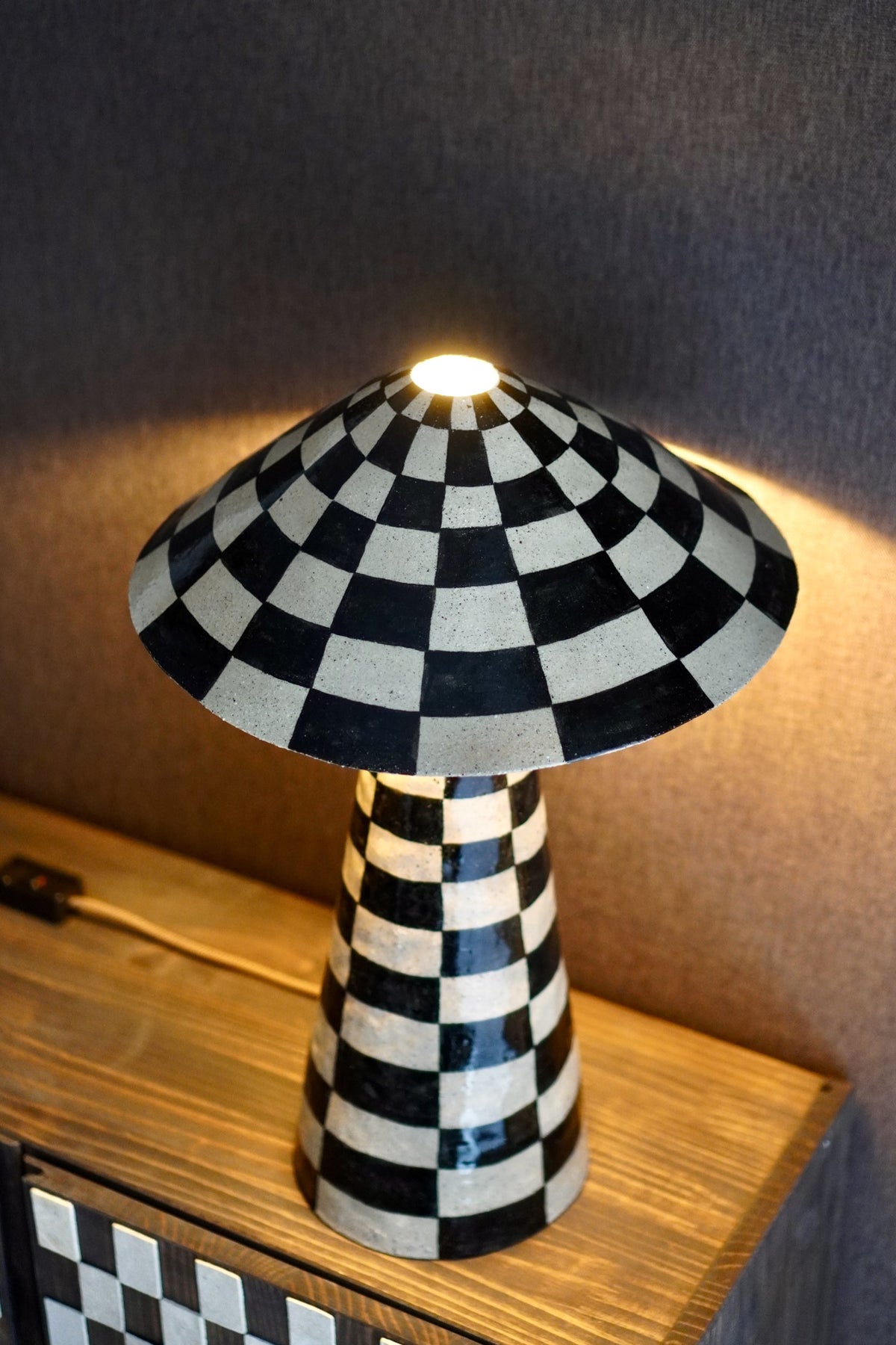 Charlie Table Lamp by Tantri Mustika, lit, its black and white checked shade glowing on a timber side table