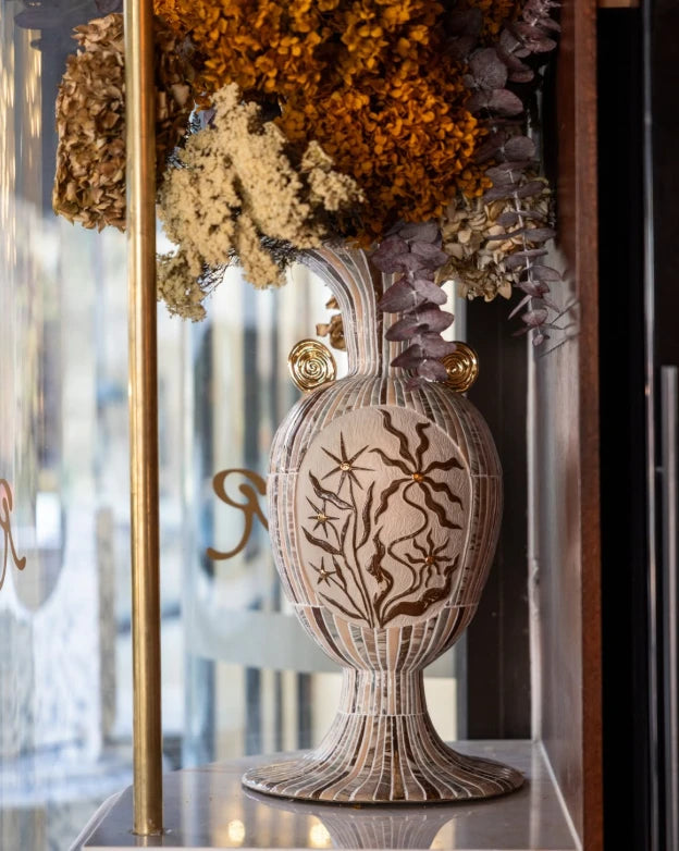 A tall vase by Tantri Mustika, striped in cream and brown, with gold spiral handles and a carved flower panel, holding dried flowers on a marble shelf