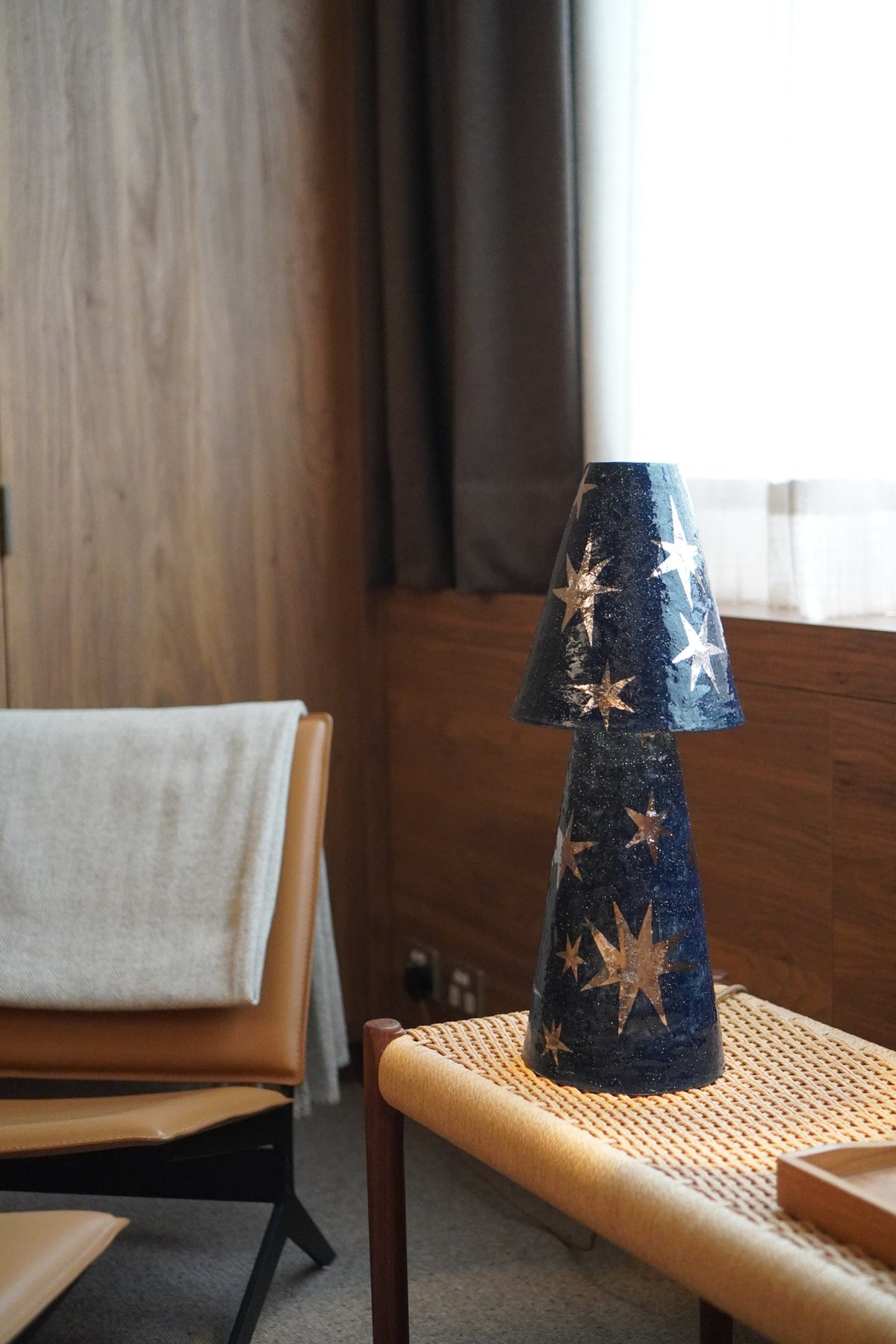 Merlin Table Lamp by Tantri Mustika on a woven bench beside a leather chair